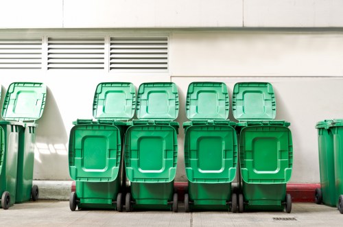 A row of eight green wheeled rubbish bins with closed lids, made of durable plastic with textured surfaces, positioned outside a building on a concrete pavement. The bins are aligned in front of a white wall with ventilation slats near the top, and a narrow red line runs along the base of the wall. The background shows even natural lighting casting soft shadows, indicating an outdoor setting. This setting reflects typical waste collection points relevant for commercial rubbish removal services provided by Office Clearance Brockley, catering to the Brockley area, which is part of South East London. The bins are clean and appear ready for waste disposal or collection, emphasizing practical waste management outside a commercial or residential property.