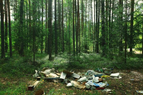 A patch of forested woodland with tall, slender trees covered in bright green leaves, allowing dappled sunlight to filter through the canopy onto the forest floor. In the foreground, there is a noticeable pile of various types of rubbish and waste materials, including cardboard boxes, plastic bags, and discarded debris, lying on the ground amidst the natural setting. The waste appears to be haphazardly strewn, contrasting with the dense, lush greenery of the trees and undergrowth. The scene suggests an area affected by illegal dumping or littering, which could be an issue for local environmental management services such as those offered by Office Clearance Brockley. The overall composition highlights the impact of improper waste disposal on natural environments near Brockley, with the background showcasing a peaceful yet disturbed woodland scene, emphasizing the need for professional rubbish removal and environmental care in the area.