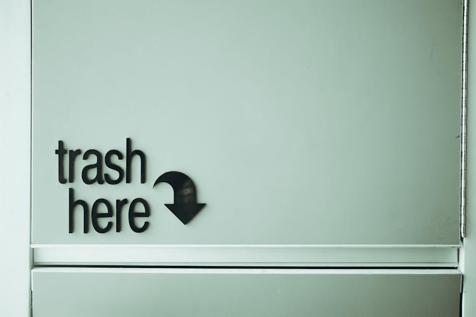 A large green wheelie bin with a white label, positioned on a paved surface beside a black brick wall. The bin is filled with red bags of garden or building waste, partially spilling over the top. The