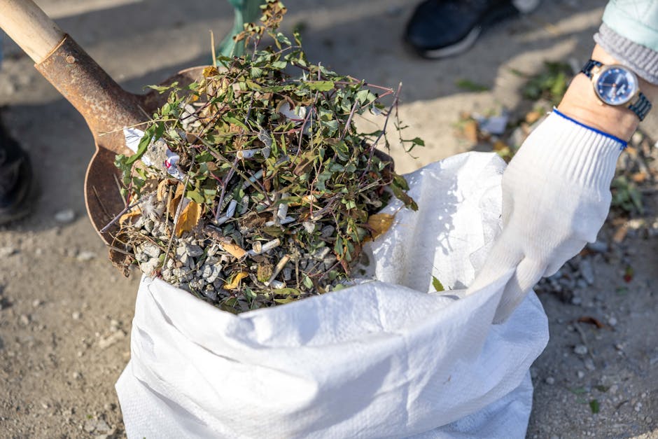 On a narrow urban pavement in front of a weathered brick and concrete building facade, two large black waste bins with hinged lids are positioned side by side, containing waste materials. One bin feat