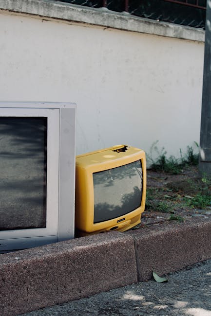 A large indoor pile of assorted electronic waste, including old CRT monitors, outdated computer towers, and vintage televisions, all haphazardly stacked against a wooden wall. The monitors and televis