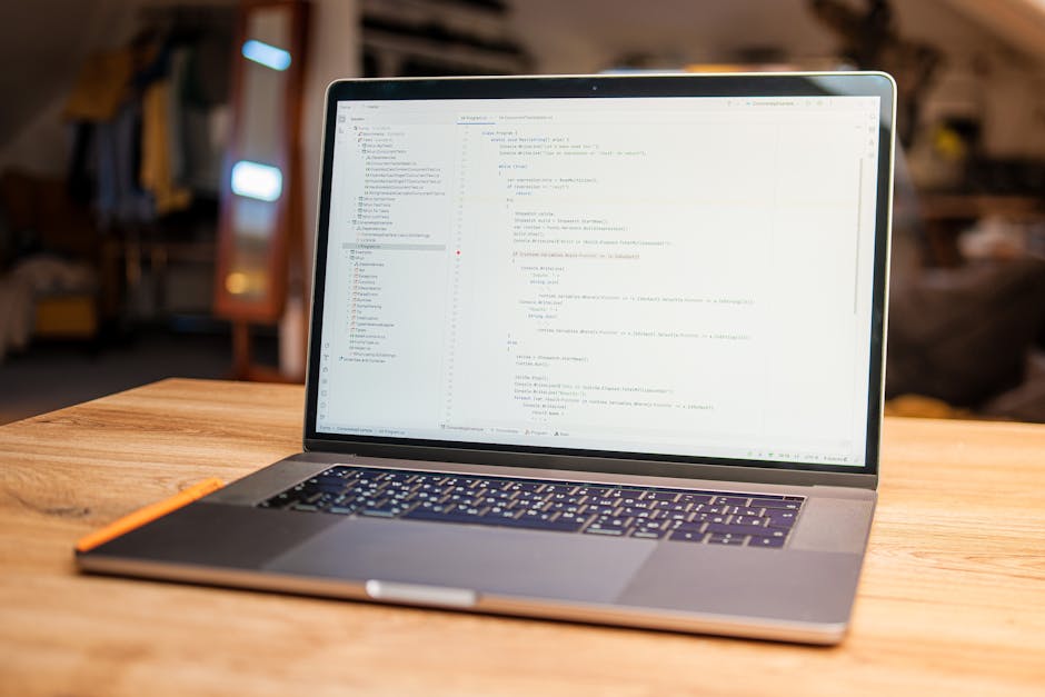 A silver laptop with a black keyboard is placed on a wooden table, displaying lines of computer code on its screen. The surface of the laptop has a smooth, metallic finish, and a slim orange stylus re