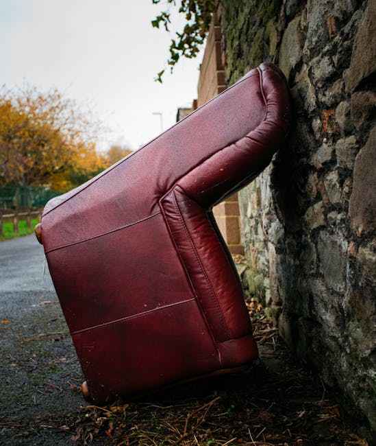A large, beige upholstered armchair with visible signs of wear and tear, including a torn section on the backrest revealing yellowish foam padding. The fabric has a slightly dirty appearance with crea